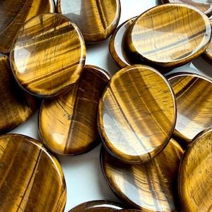 May include: A collection of polished tiger's eye stones. The stones are oval and round, with a golden-brown color and chatoyant bands. The stones are arranged on a white surface, with some overlapping. The tiger's eye stones are a natural gemstone.