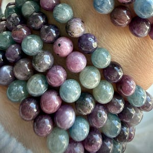 May include: A multi-strand bracelet composed of polished, spherical beads in varying shades of purple, pink, and light blue. The beads feature natural inclusions and subtle color variations, arranged in a repeating pattern along the elastic cord.