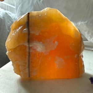May include: A large, raw, orange calcite crystal with a translucent appearance. The crystal has a rough, uneven surface and a warm, glowing interior. A dark brown line runs vertically through the center. The crystal is displayed on a white surface.