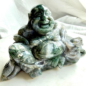 May include: A carved stone figurine of a laughing Buddha, crafted from moss agate with deep green and translucent grey inclusions. The sculpture depicts a seated figure with a prominent belly, a wide smiling face, and smooth, polished surfaces.
