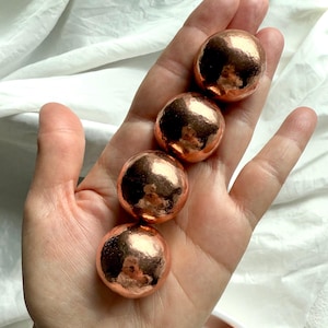 May include: Five smooth, copper spheres, each with a slightly different shade of copper, are held in a hand. The spheres are about the size of a golf ball.