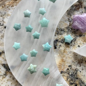 May include: A white crescent moon-shaped selenite crystal with a variety of green star-shaped crystals arranged on the surface. A single purple star-shaped crystal is placed on the top of the moon.