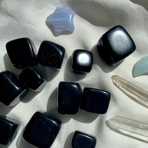 May include: A collection of black tumbled stones, also known as black obsidian, on a white fabric background. There are 12 stones in total, with a few smaller stones and a few larger stones. There is also a blue star-shaped stone and a clear quartz crystal.