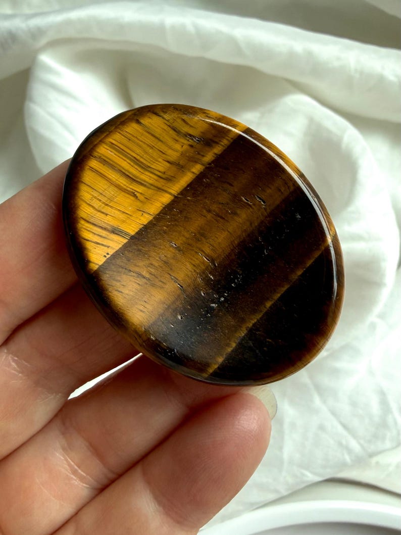 close-up tiger’s eye worry stone in hand with strong chatoyancy flash, grounding crystal for solar plexus confidence and clarity