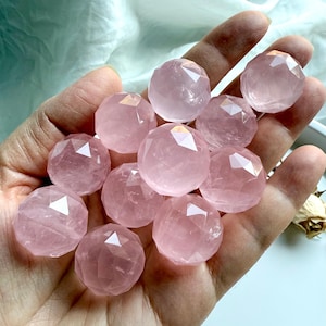 May include: A collection of faceted, spherical rose quartz crystals. The pale pink stones are held in a hand, showcasing their geometric cuts and translucent quality. The crystals are a soft, natural pink color.
