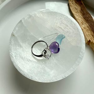 May include: A white selenite bowl holds a silver ring with a clear gemstone, a purple heart-shaped crystal, a blue crystal, and a clear crystal wand. A piece of wood rests on the side of the bowl.