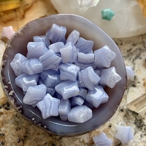 May include: A collection of small, light blue star-shaped crystals fills a gray and brown agate bowl. The crystals have a layered, marbled appearance. Additional star-shaped crystals are scattered around the bowl, with a few more in the background.