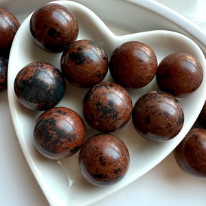 May include: A collection of polished, spherical gemstones in shades of brown and black, displayed in a white heart-shaped dish. The stones have a glossy finish and varying patterns.