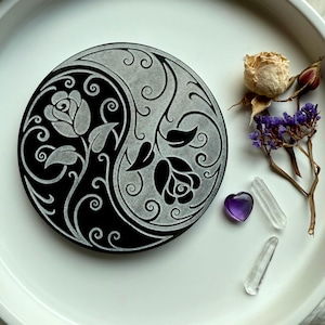 May include: A black and silver Yin Yang design with floral accents. The circular design features roses and swirling patterns. A dried rose, purple flowers, a heart-shaped amethyst, and clear quartz crystals are arranged nearby on a white plate.
