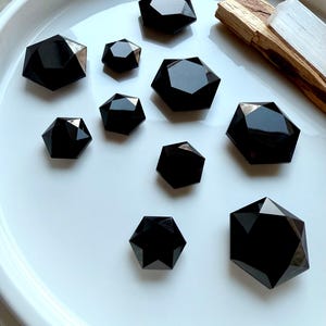 Faceted Black Obsidian hexagon crystals in assorted sizes displayed on a white dish with Palo Santo and Selenite — grounding root chakra stones for protection and energy clearing.