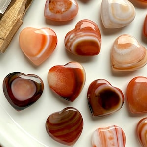 May include: A collection of heart-shaped stones in shades of orange, brown, and white. The polished stones have a glossy finish and varying patterns, displayed on a white surface. These decorative stones are ideal for gifts or home decor.