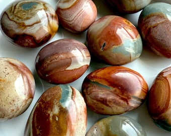 May include: A selection of polished stones in shades of brown, red, cream, and blue-green, presented on a white plate. The stones have a smooth, glossy finish and diverse patterns. These decorative stones are suitable for home decor or as a unique gift.