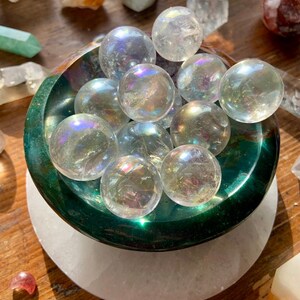 May include: A green bowl filled with iridescent clear quartz spheres. The bowl is sitting on a white circular base.