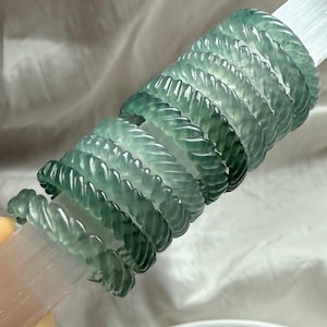 May include: A collection of jade rings in varying shades of green, arranged on a clear, cylindrical object. The rings have a twisted rope design, and the overall aesthetic is elegant and natural. The rings are a beautiful accessory.