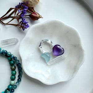 White marble daisy bowl with scalloped flower edges holding an amethyst heart carving, aquamarine moon, and a clear quartz hexagon