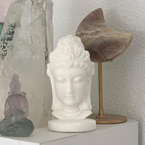 White Jade Quan Yin Statue shown on a shelf with some larger crystals and another Quan Yin figurine in the picture.