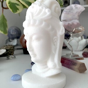 White Jade Quan Yin carving shown on a white table with other crystals and a plant in the background