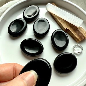 May include: A collection of polished black obsidian stones in an oval shape, displayed on a white plate. A white selenite wand and a piece of palo santo are also present. A hand is holding one of the obsidian stones. A clear crystal is also present.