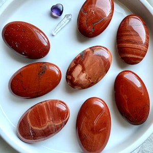 May include: A collection of polished red jasper stones in various oval shapes, displayed on a white ceramic plate. The stones have unique patterns and textures, with a small purple heart-shaped stone and a clear crystal point also present.