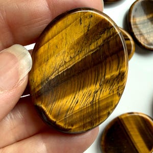 tiger’s eye worry stone held between fingers showing natural banding and sheen, crystal for protection strength and balance
