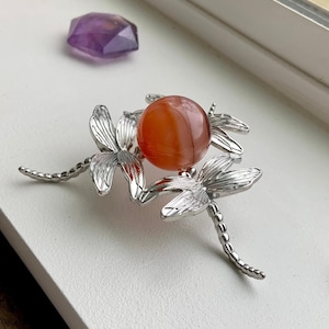 May include: A silver dragonfly design with three dragonflies holding a round orange carnelian stone in the center.