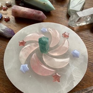 Crystal grid spiral of rose quartz moons, blue lace agate stars, and strawberry quartz stars on selenite plate with other crystals on dark wood table