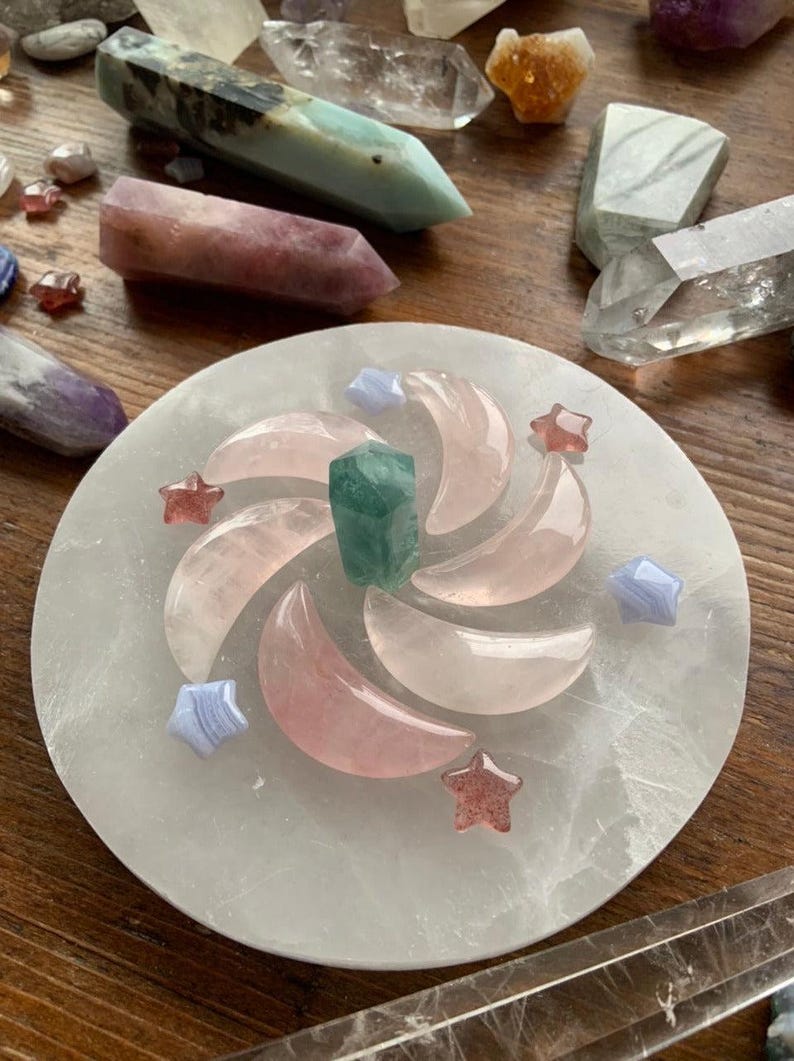 Crystal grid spiral of rose quartz moons, blue lace agate stars, and strawberry quartz stars on selenite plate with other crystals on dark wood table
