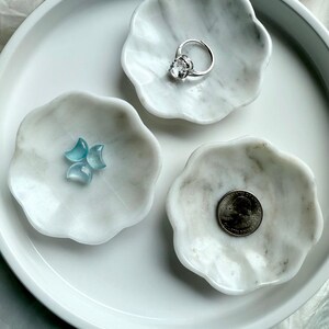 Three white marble daisy bowls with natural gray veining, one styled with a silver ring, one with aquamarine crescent carvings, and one with a quarter for size reference