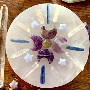Overhead of crystal grid on selenite plate with kyanite sticks, clear quartz points, amethyst moons, and blue lace stars on dark wood altar table