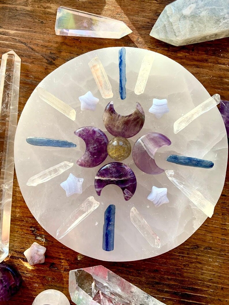 Overhead of crystal grid on selenite plate with kyanite sticks, clear quartz points, amethyst moons, and blue lace stars on dark wood altar table