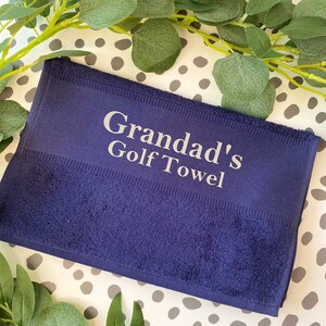 May include: A navy blue golf towel with the text "Grandad's Golf Towel" in white. The towel is rectangular and has a textured surface. Green leaves and a dotted background surround the towel.