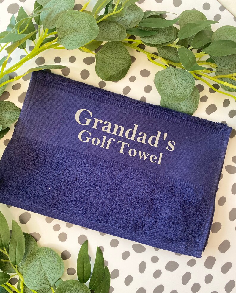 May include: A navy blue golf towel with the text "Grandad's Golf Towel" in white. The towel is rectangular and has a textured surface. Green leaves and a dotted background surround the towel.