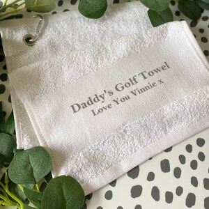 May include: White golf towel with the text "Daddy's Golf Towel Love You Vinnie x" in gray. The towel has a metal grommet and a loop for hanging. Greenery is arranged around the towel on a white and gray spotted surface.