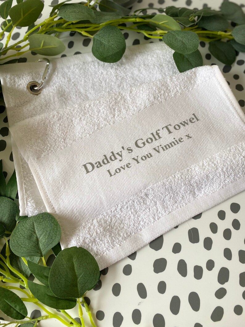 May include: White golf towel with the text "Daddy's Golf Towel Love You Vinnie x" in gray. The towel has a metal grommet and a loop for hanging. Greenery is arranged around the towel on a white and gray spotted surface.