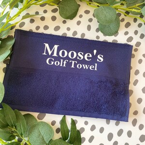 May include: A navy blue golf towel with the text "Moose's Golf Towel" in white. The towel is rectangular and has a textured surface. Green leafy branches are arranged around the towel on a white and gray polka dot background.