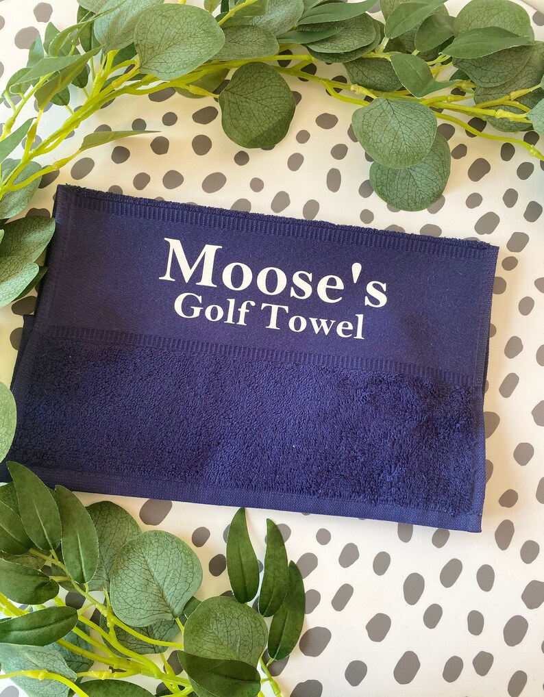 May include: A navy blue golf towel with the text "Moose's Golf Towel" in white. The towel is rectangular and has a textured surface. Green leafy branches are arranged around the towel on a white and gray polka dot background.