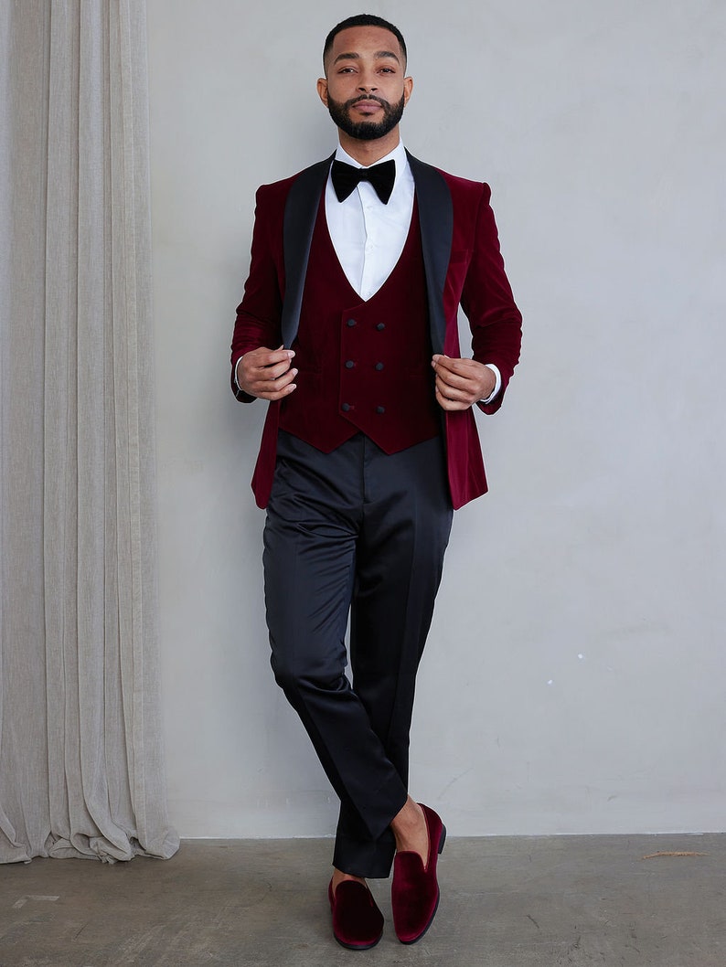 Pu&ograve; includere: Un abito in velluto bordeaux con revers in raso nero e un gilet in velluto bordeaux abbinato. L'abito &egrave; indossato con una camicia bianca e un papillon nero. Il modello indossa mocassini in velluto bordeaux.