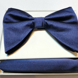 Puede incluir: Un conjunto de pajarita y pañuelo de bolsillo azul marino. La pajarita está preanudada y el pañuelo de bolsillo está hecho de una tela sedosa.