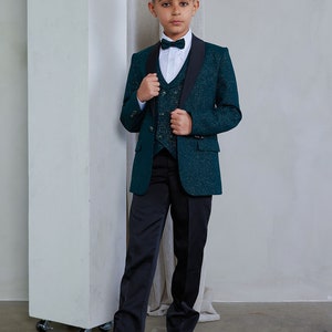 Op de afbeelding: Een jonge jongen draagt een donkergroen pak met een zwarte strik en vest. Het pak heeft een subtiele glans in de hele stof. De jongen staat voor een witte muur.