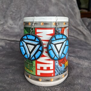 May include: A pair of blue and silver earrings with a circular design featuring the Iron Man symbol. The earrings are hanging from a mug with a Marvel logo.