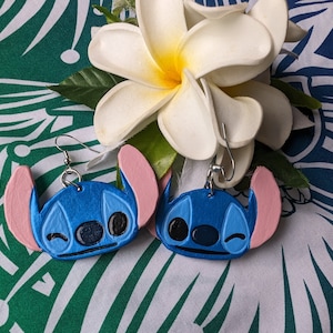May include: A pair of blue and pink Stitch earrings. The earrings are made of polymer clay and feature a winking Stitch face.