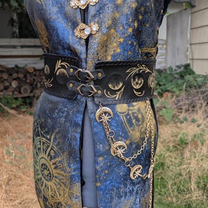 Op de afbeelding: Een blauw en goud hemels thema vest met een zwarte leren riem. De riem heeft gouden maan- en sterontwerpen en is vastgemaakt met twee gespen. Een gouden ketting met maan- en sterbedels hangt aan de riem.