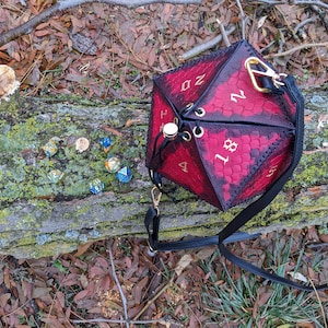 May include: A red and black D20 dice-shaped bag with gold details and a black strap. The bag has a dragon scale design. Several dice are scattered around the bag on a log.