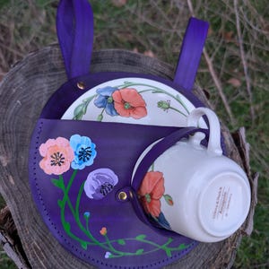 May include: A purple leather tea cup holder with a matching handle, holding a white teacup and a floral plate. The holder is hand-painted with colorful poppy flowers and green vines.