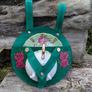 Può includere: Un porta-set da tè in pelle verde smeraldo fatto a mano con una teiera bianca e un piattino decorato con fiori. Il porta-set presenta un motivo a rosa e rivetti color oro. Il porta-set ha due cinghie verdi per il trasporto.
