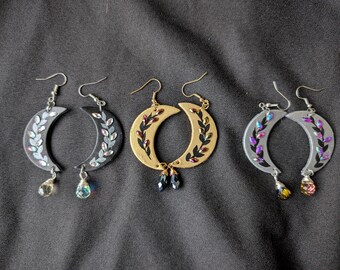 May include: Three pairs of crescent moon earrings. One pair is silver with clear and iridescent teardrop-shaped charms. Another pair is gold with black and blue charms. The last pair is silver with purple and yellow charms.