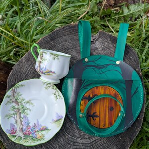Può includere: Un set di tazza e piattino bianchi con un disegno di albero e fiori e un bordo verde. Accanto, una borsa in pelle verde e nera con un disegno di porta in legno.
