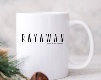 Puede incluir: Una taza de cerámica blanca con el texto negro BAYAWAN y el texto más pequeño AGUSAN DEL SUR, PHILIPPINES impresos en el frente. La taza tiene un acabado liso, un cuerpo cilíndrico y un asa curva en el lado derecho para sujetarla.