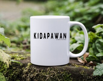 Puede incluir: Una taza de cerámica blanca con el texto negro KIDAPAWAN y el texto más pequeño COTABATO, PHILIPPINES impresos en el frente. La taza tiene un cuerpo cilíndrico liso y un asa curva en el lado derecho para sostener bebidas calientes.