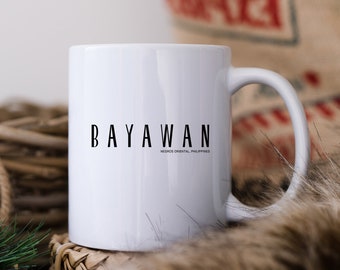 Puede incluir: Una taza de cerámica blanca con el texto negro BAYAWAN en una fuente alta sin remates, con el texto más pequeño NEGROS ORIENTAL, PHILIPPINES debajo. La taza tiene un acabado liso y brillante con un asa curva en el lado derecho.